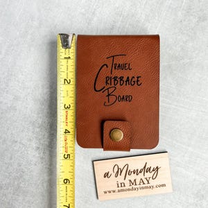 以下が含まれることがあります： 前面に「TRAVEL CRIBBAGE BOARD」と刻まれた茶色の革製の旅行用クリbbageボード。手前に「a Monday in MAY www.amondayinmay.com」と書かれた小さな木製の看板がある。