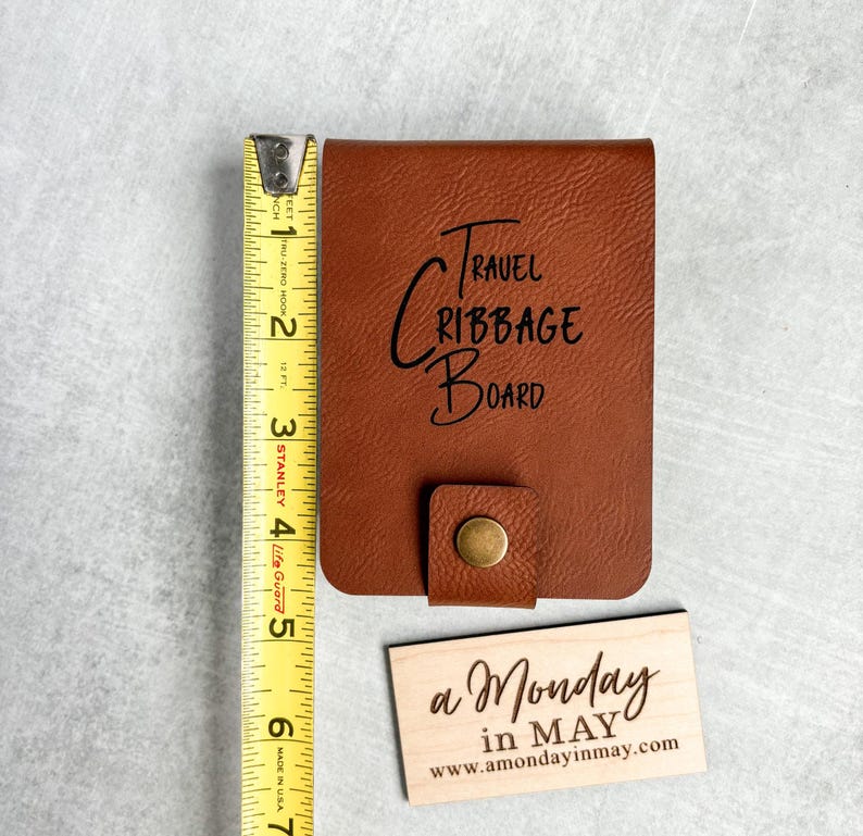 以下が含まれることがあります： 前面に「TRAVEL CRIBBAGE BOARD」と刻まれた茶色の革製の旅行用クリbbageボード。手前に「a Monday in MAY www.amondayinmay.com」と書かれた小さな木製の看板がある。