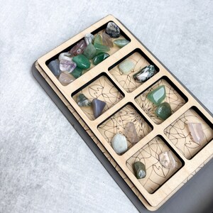 May include: A wooden tray with nine compartments, each containing a different type of stone. The stones are various shades of green, pink, and gray. The tray is decorated with a mandala design.