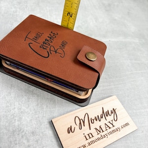 以下が含まれることがあります： 前面に「Travel Cribbage Board」と刻まれた茶色の革張りの旅行用クリbbageボード。ボードは閉じており、その背後にはメジャーが一部見えている。手前に「a Monday in MAY www.amondayinmay.com」と書かれた木製の看板がある。