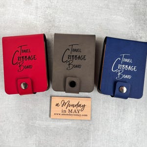 May include: Three travel cribbage boards in red, brown, and blue leather. Each board has the text "Travel Cribbage Board" printed on the front.