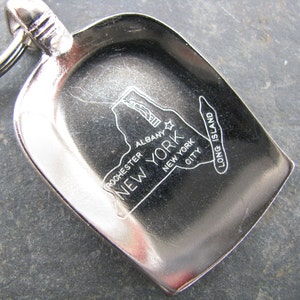 May include: A silver-colored keychain featuring a shovel-shaped charm. The charm has an engraved map of New York State, with the cities of Albany, Rochester, and New York City labeled. The words "Long Island" are also visible.