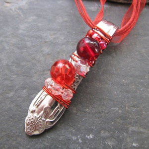 May include: A handmade necklace featuring a repurposed silver spoon pendant. The spoon is adorned with red beads and wire wrapping, suspended from a red ribbon. The pendant has a floral design on the handle.