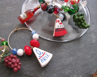 May include: Wine glass charms with gold hoops, featuring red wine bottle, cheese wedge, and grape cluster charms. The charms are accented with colorful beads in red, white, green, and black. The wine glass is partially visible.
