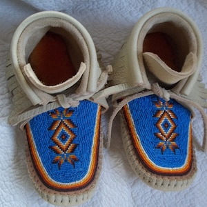 May include: A pair of cream-colored leather moccasins with blue beaded designs. The beaded section features a central diamond shape with orange and brown accents. The moccasins have fringe detailing and leather laces.