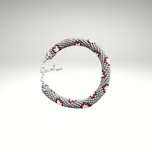 Pode incluir: Um bracelete cinzento de contas com detalhes vermelhos e brancos. O bracelete é feito de contas pequenas e tem um fecho prateado.
