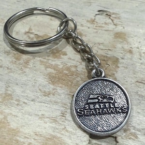 May include: Silver-colored Seattle Seahawks keychain. The round pendant features the team logo and the words "Seattle Seahawks" in a textured, metallic finish. The keychain includes a chain and a ring.
