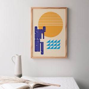 Puede incluir: Una impresi&oacute;n enmarcada con un dise&ntilde;o retro que presenta un sol amarillo con rayas naranjas, olas azules y el texto "Surf City" en azul.
