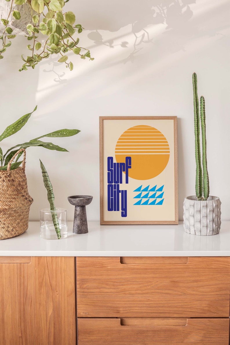 Puede incluir: Una impresi&oacute;n enmarcada con un dise&ntilde;o retro que presenta un sol amarillo con rayas naranjas, olas azules y el texto "Surf City" en azul.