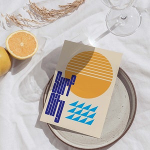 Puede incluir: Una tarjeta beige con un sol amarillo y tri&aacute;ngulos azules. El texto "Surf City" est&aacute; escrito en azul en la tarjeta.