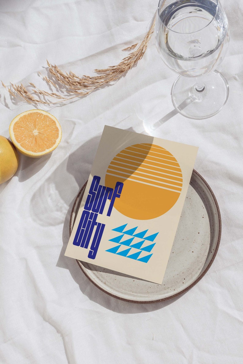 Puede incluir: Una tarjeta beige con un sol amarillo y tri&aacute;ngulos azules. El texto "Surf City" est&aacute; escrito en azul en la tarjeta.