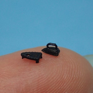 Puede incluir: Dos objetos de hierro en miniatura de color negro, una pequeña plancha y una pequeña tabla de planchar, se muestran en un dedo.