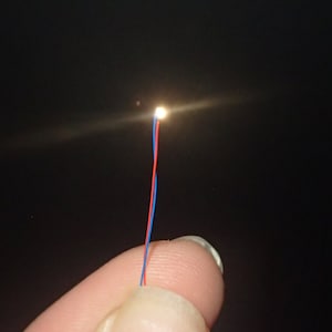 Op de afbeelding: Een klein, warm wit LED-lampje met rode en blauwe draden eraan bevestigd. Het lampje wordt vastgehouden door een vinger.