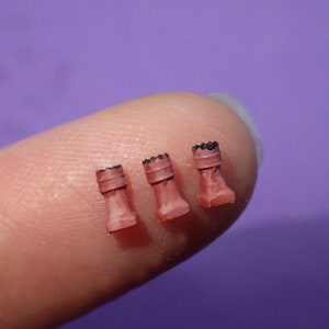 May include: Three miniature, reddish-pink rook chess pieces with black crenellations. The tiny figurines are arranged on a fingertip against a purple background. The rook pieces are detailed with a cylindrical body and a square base.