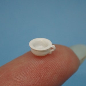May include: A miniature white porcelain chamber pot with a handle. The pot is small enough to fit on a fingertip.