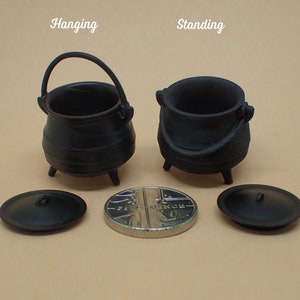 Puede incluir: Dos ollas de hierro fundido negro, una colgada y otra de pie, con tapas. Las ollas son del tamaño de una taza de té. Una moneda británica está en primer plano para la escala.