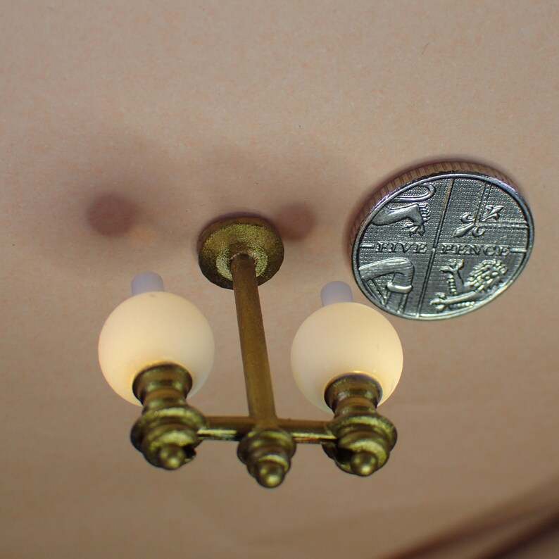 May include: A gold-toned, two-light chandelier with white glass shades. The chandelier is hanging from a ceiling and is about the size of a British five pence coin.