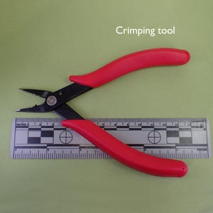 May include: Red and black crimping tool with a ruler in the background. The tool is used for crimping jewelry wire.