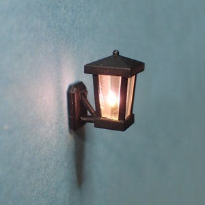 Puede incluir: Un aplique de pared negro con una pantalla de vidrio transparente y una bombilla de luz blanca cálida. El aplique está montado en una pared blanca.