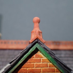 May include: A close-up of a miniature brick building with a green and black roof and a terracotta chimney. The building has a red brick facade and a green trim.