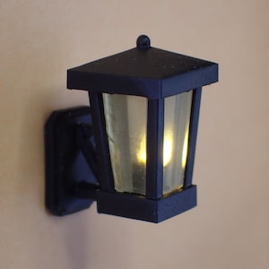 Può includere: Lanterna da parete nera con un pannello in vetro trasparente e una luce LED bianco caldo all'interno.