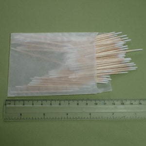 Könnte beinhalten: Eine Packung Holz-Wattestäbchen mit weißen Spitzen, teilweise sichtbar in einer durchscheinenden Papierverpackung. Ein transparentes Lineal mit Zentimetermarkierungen liegt darunter und misst bis zu 15 cm.