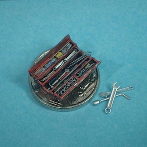 May include: A miniature red toolbox containing various tools, including spanners and screwdrivers. The toolbox is open, displaying the tools neatly arranged. Several silver spanners are placed next to the toolbox. The items are on a silver coin.