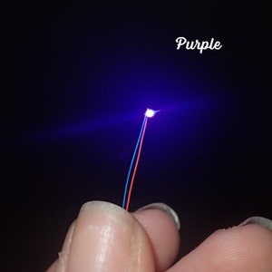 Op de afbeelding: Een klein LED-lampje dat een helder paars licht uitstraalt. Het lampje is bevestigd aan twee draden, een rode en een blauwe.