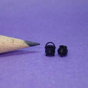 Puede incluir: Dos pequeños calderos de metal negro con asas, junto a un lápiz sobre una superficie morada.