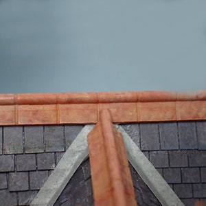 May include: A close-up view of a roof with a terracotta tile ridge and a slate roof. The roof is angled and has a grey concrete trim.