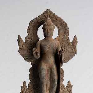 Könnte beinhalten: Eine Bronzestatue eines stehenden Buddha mit einem detaillierten, verzierten Sockel und einem Heiligenschein hinter der Figur. Die Statue ist in einem verwitterten, gealterten Zustand.
