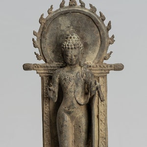 Op de afbeelding: Een bronzen beeld van een staande Boeddha, met een halo achter het hoofd, en een kleinere Boeddha die eronder zit. Het beeld staat op een sokkel met een decoratieve basis.