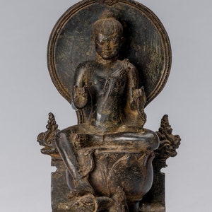 Op de afbeelding: Een donker bronzen beeld van een zittend Boeddhabeeld met een halo achter het hoofd. De Boeddha draagt een gewaad en heeft een serene uitdrukking. Het beeld zit op een lotusthron met een basis versierd met bloemmotieven.