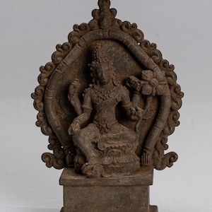 Può includere: Una scultura in metallo marrone scuro di una divinità seduta con quattro braccia, che tiene un fiore di loto in una mano. La scultura è posta su una base quadrata ed è circondata da una cornice decorativa.