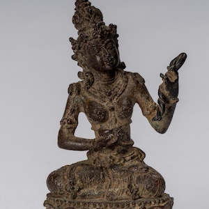 Può includere: Una statua di bronzo di una divinità seduta con una corona e gioielli elaborati. La divinità tiene un piccolo oggetto nella mano destra e ha un'espressione serena.