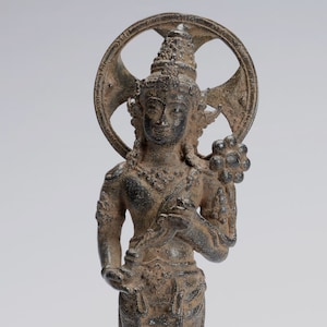May include: A dark grey bronze statue of a standing figure with a halo and a lotus flower in their hand. The figure is wearing a long robe and a crown. The statue is on a pedestal with a lotus flower design.