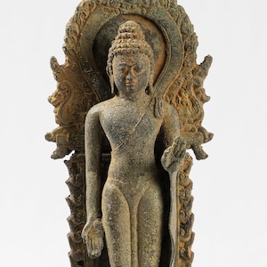 Op de afbeelding: Een bronzen beeld van een staande Boeddha, met een halo en ingewikkelde details, op een voetstuk met een decoratief ontwerp.