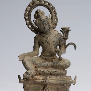 Può includere: Statua color bronzo di una figura seduta con un'aureola, adornata con dettagli intricati. La figura è seduta su un piedistallo decorativo. La statua presenta un'espressione serena e tiene in mano un oggetto a forma di loto.