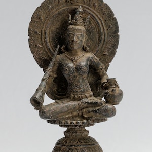 Può includere: Una statua di bronzo di una divinità seduta con un'aureola e uno strumento musicale. La divinità tiene una pentola in una mano e uno strumento musicale nell'altra. La statua è su un piedistallo con una base.