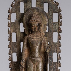 May include: A bronze statue of a standing Buddha, with a halo and a patterned robe, set within a decorative frame. The statue is on a base with intricate carvings.