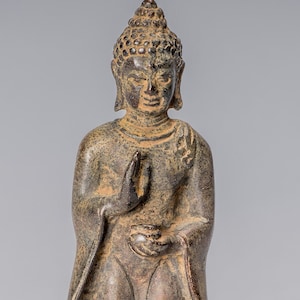 Puede incluir: Una estatua de bronce de un Buda de pie con un tocado tradicional y una expresión serena. El Buda lleva una túnica larga y tiene una flor de loto en la mano. La estatua está sobre una base circular con tallas intrincadas.