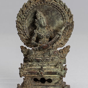 Può includere: Una scultura in bronzo di una figura seduta che suona uno strumento a corda. La figura è circondata da un'aureola circolare e siede su un trono. La scultura è patinata di verde.