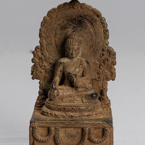 Puede incluir: Una estatua de bronce de un Buda sentado, con una corona puntiaguda y un halo circular detrás de la figura. El Buda está sentado en un trono de loto con una base decorativa.