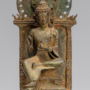 Pode incluir: Uma estátua de bronze de um Buda sentado com um halo e uma base decorativa. A estátua está em uma postura meditativa com uma mão levantada em um gesto de bênção.