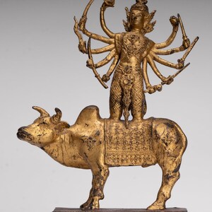 Peut inclure: Statue dorée représentant une divinité à plusieurs bras chevauchant un taureau. La divinité est ornée de détails complexes et tient divers objets. Le taureau est également finement détaillé, avec une selle à motifs. La statue est montée sur une base rectangulaire.
