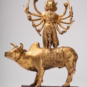 Può includere: Una statua dorata raffigurante una divinità con molte braccia in piedi su un toro dorato. La divinità è adornata con dettagli intricati, che tiene in mano vari oggetti. Anche il toro è molto dettagliato, con una sella decorativa. La statua è montata su una base rettangolare.