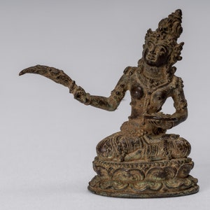 Op de afbeelding: Een bronzen beeld van een zittende godin die een gestileerd mes in de ene hand en een klein schoteltje in de andere hand houdt. De godin is versierd met uitgebreide sieraden en een kroon.