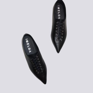 K&ouml;nnte beinhalten: Ein Paar schwarze Lederschn&uuml;rschuhe mit spitz zulaufenden Zehen. Die Schuhe haben das Wort "IMELDA" an der Seite.