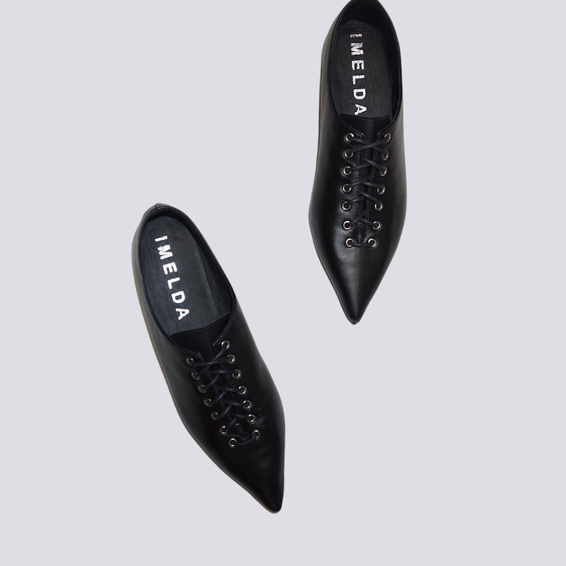 K&ouml;nnte beinhalten: Ein Paar schwarze Lederschn&uuml;rschuhe mit spitz zulaufenden Zehen. Die Schuhe haben das Wort "IMELDA" an der Seite.