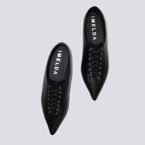 K&ouml;nnte beinhalten: Ein Paar schwarze Lederschn&uuml;rschuhe mit spitz zulaufenden Zehen. Die Schuhe tragen den Schriftzug "IMELDA" auf der Innenseite jedes Schuhs.